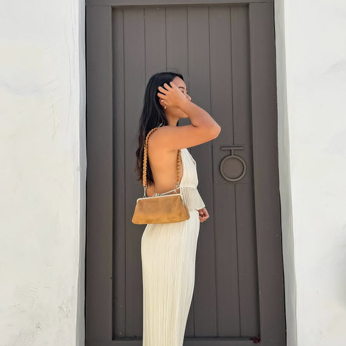 A woman in a cream pleated backless maxi dress stands in profile holding a tan leather shoulder bag with a braided strap, set against a dark gray wooden door and white walls.